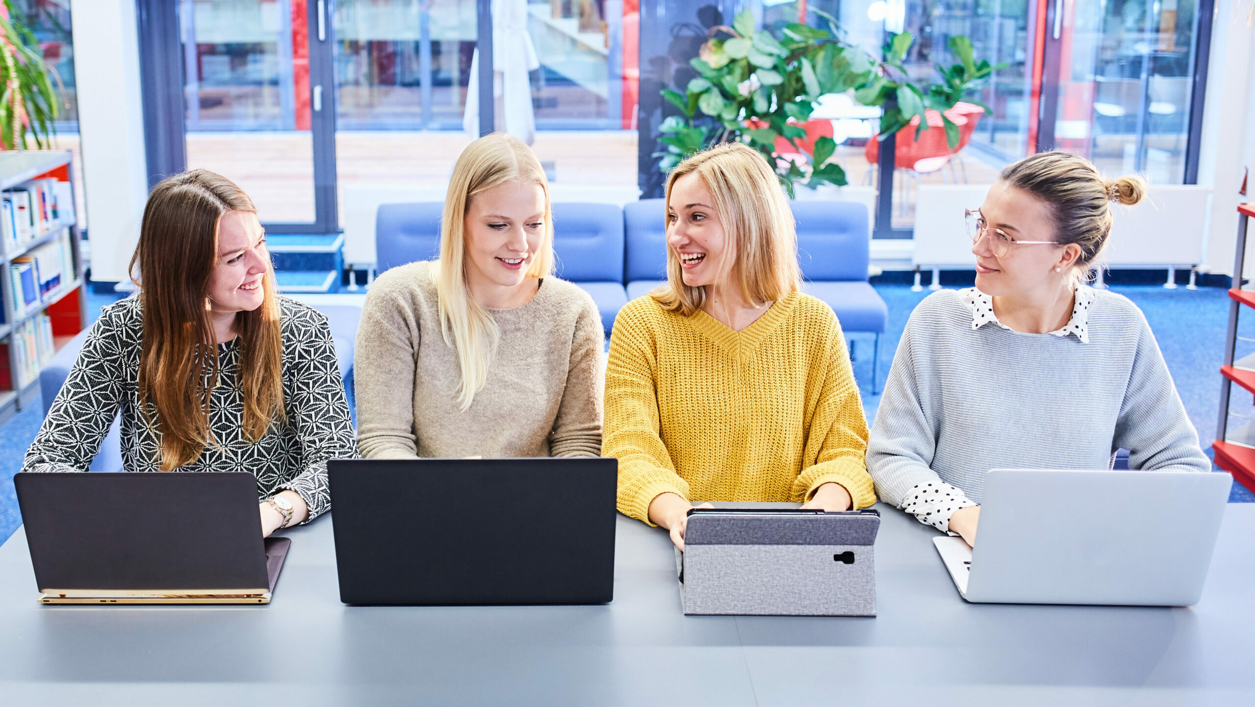 Vier Studentinnen sitzen nebeneinander und unterhalten sich vor ihren aufgeklappten Laptops in der Bibliothek der Hochschule Kehl.