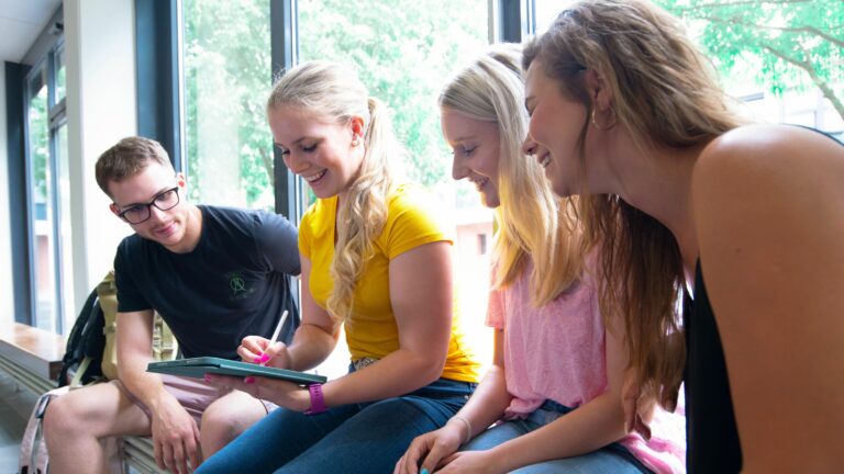 Vier Studierende sitzen nebeneinander im Foyer der Hochschule und schauen gemeinsam auf ein Tablet
